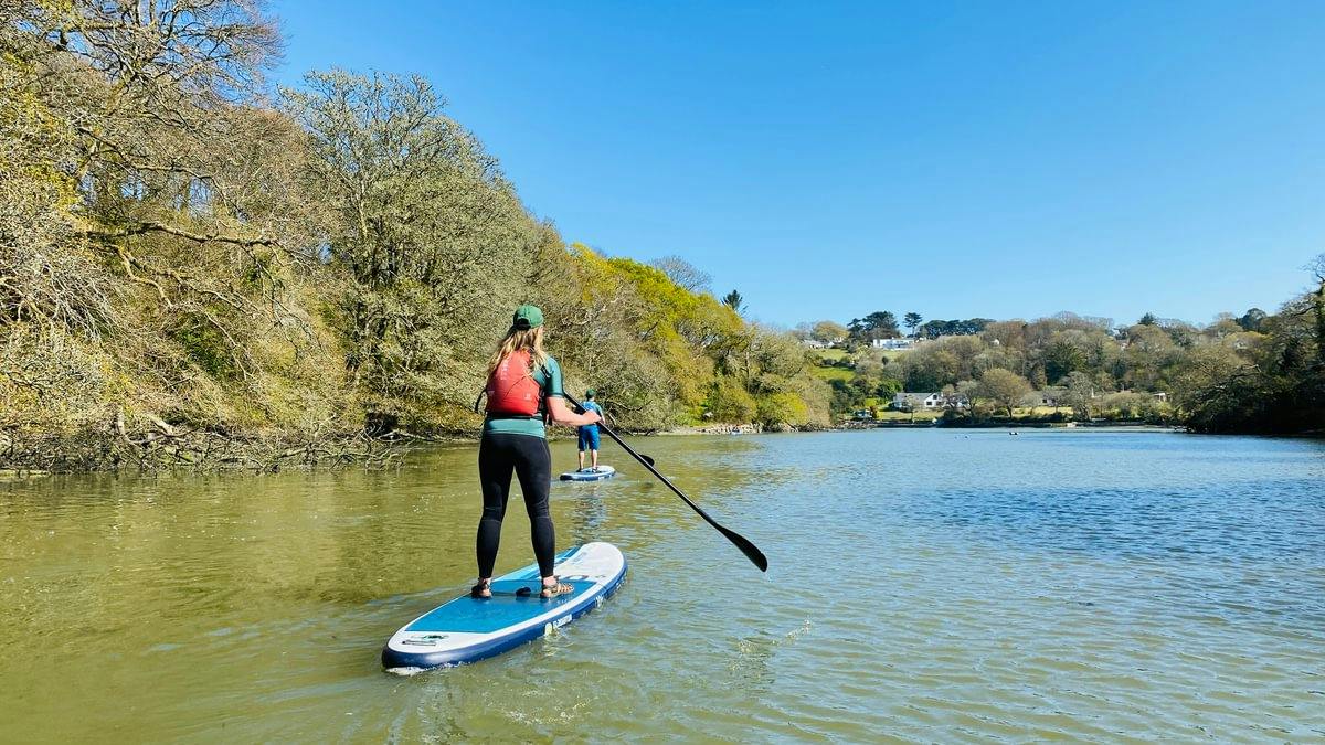 Person paddleboarding on a calm river under a clear blue sky, with another paddler ahead and trees lining both banks.