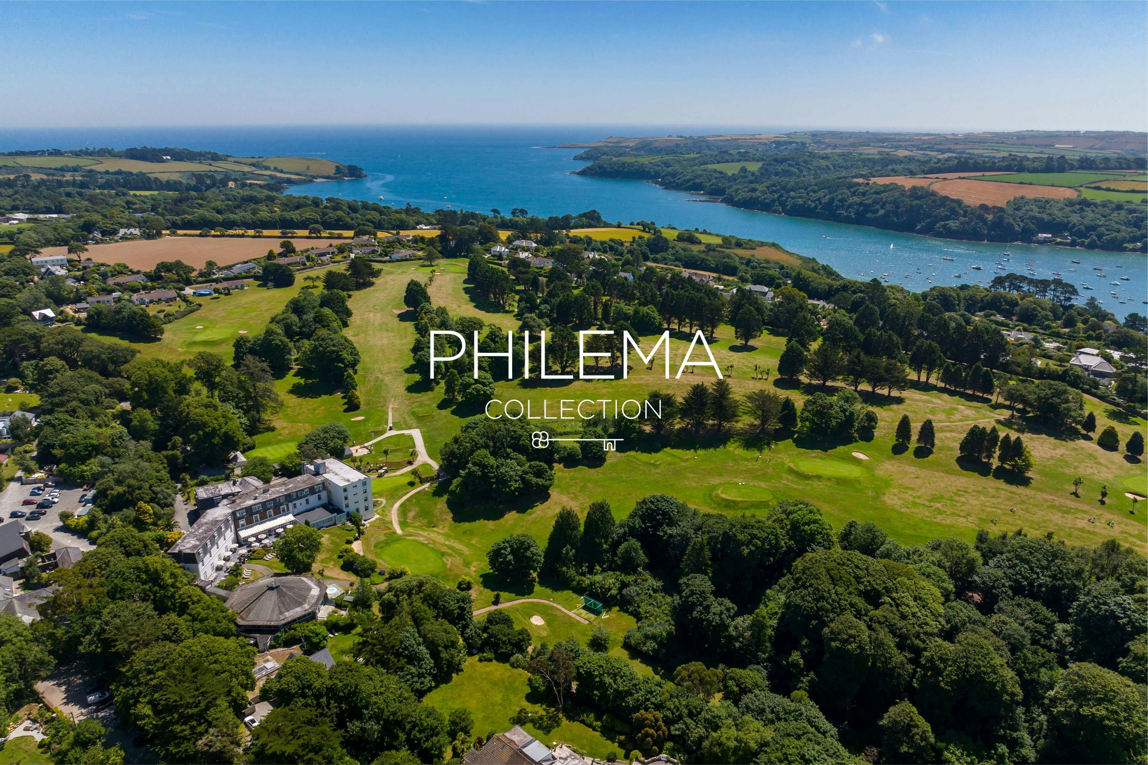 Aerial view of a coastal golf resort with hotel, green fairways, trees, and a blue estuary under clear skies.