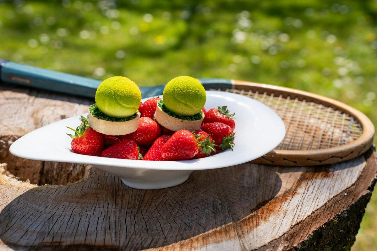 Plate of strawberries topped with two tennis ball-shaped appetizers on a tree stump, with a tennis racket beside it outdoors.