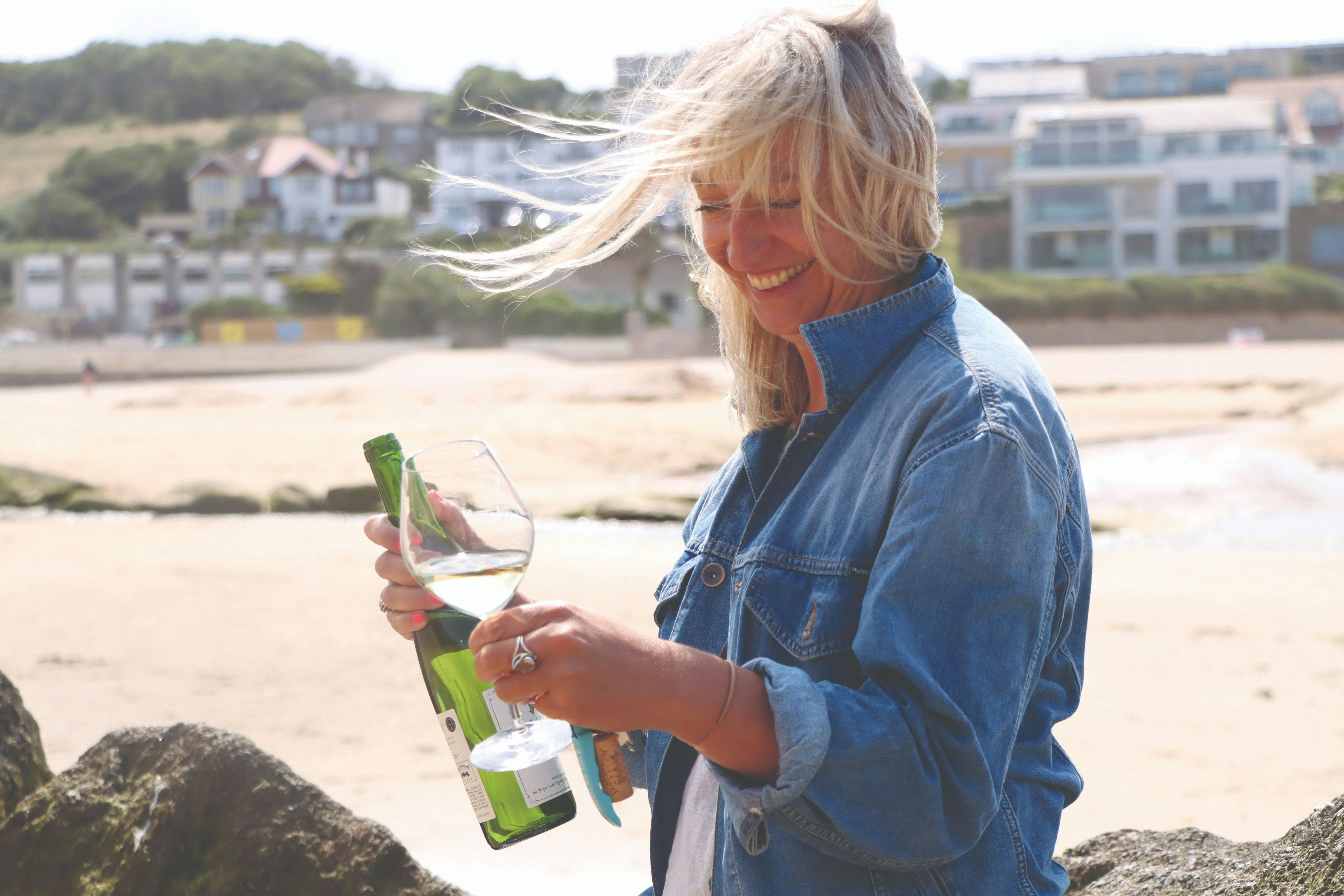 Smiling woman in a denim jacket on a windy beach, holding a wine bottle, glass, and corkscrew with houses blurred behind her.