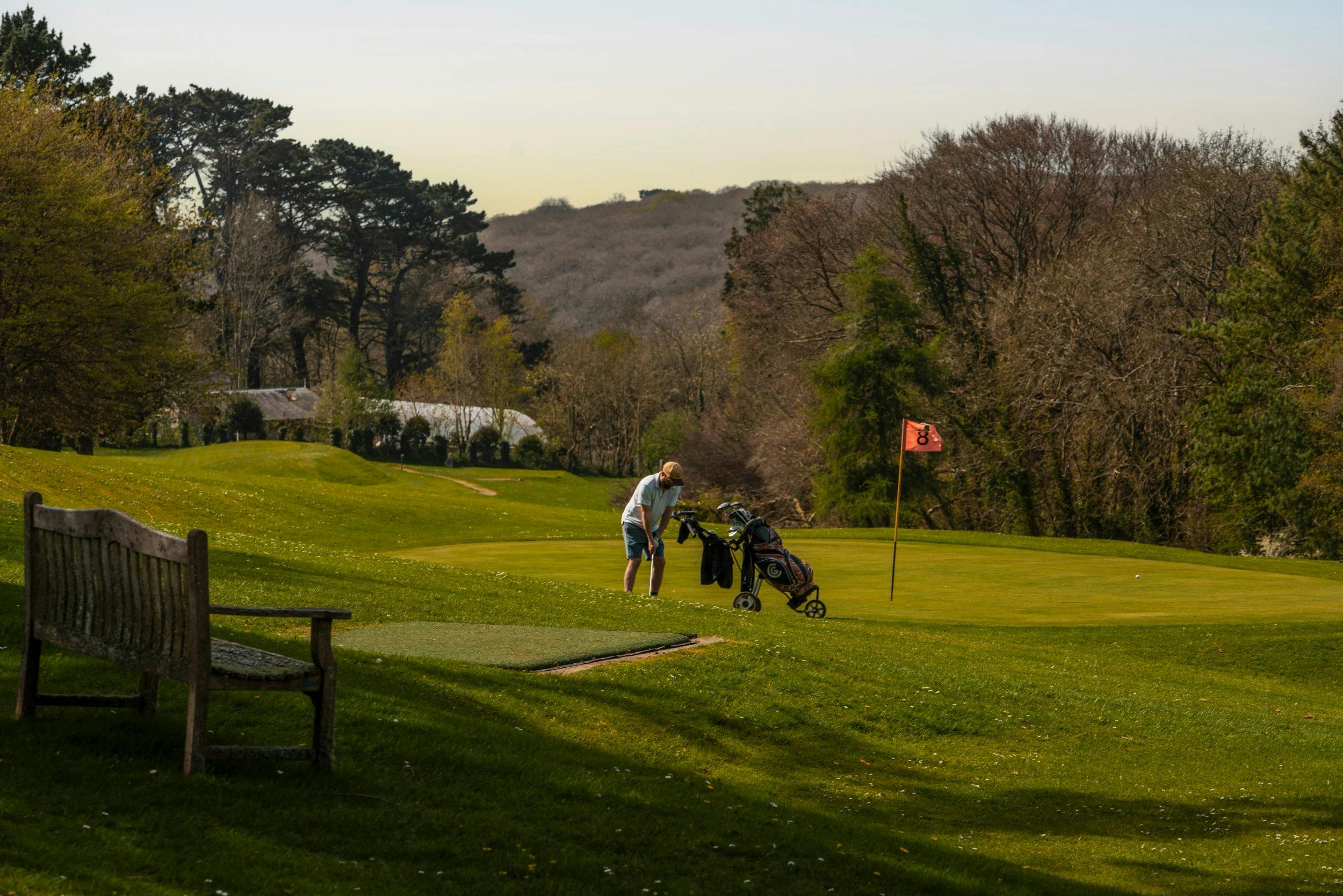 Golfer putts on a green beside an orange flag, with a golf cart, bench, and wooded hills in the background.
