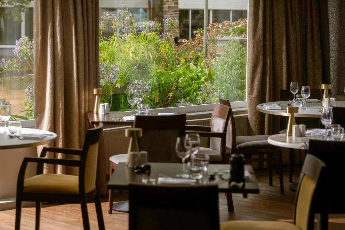 Quiet restaurant dining room with set tables, cushioned chairs, and large windows overlooking lush garden flowers.