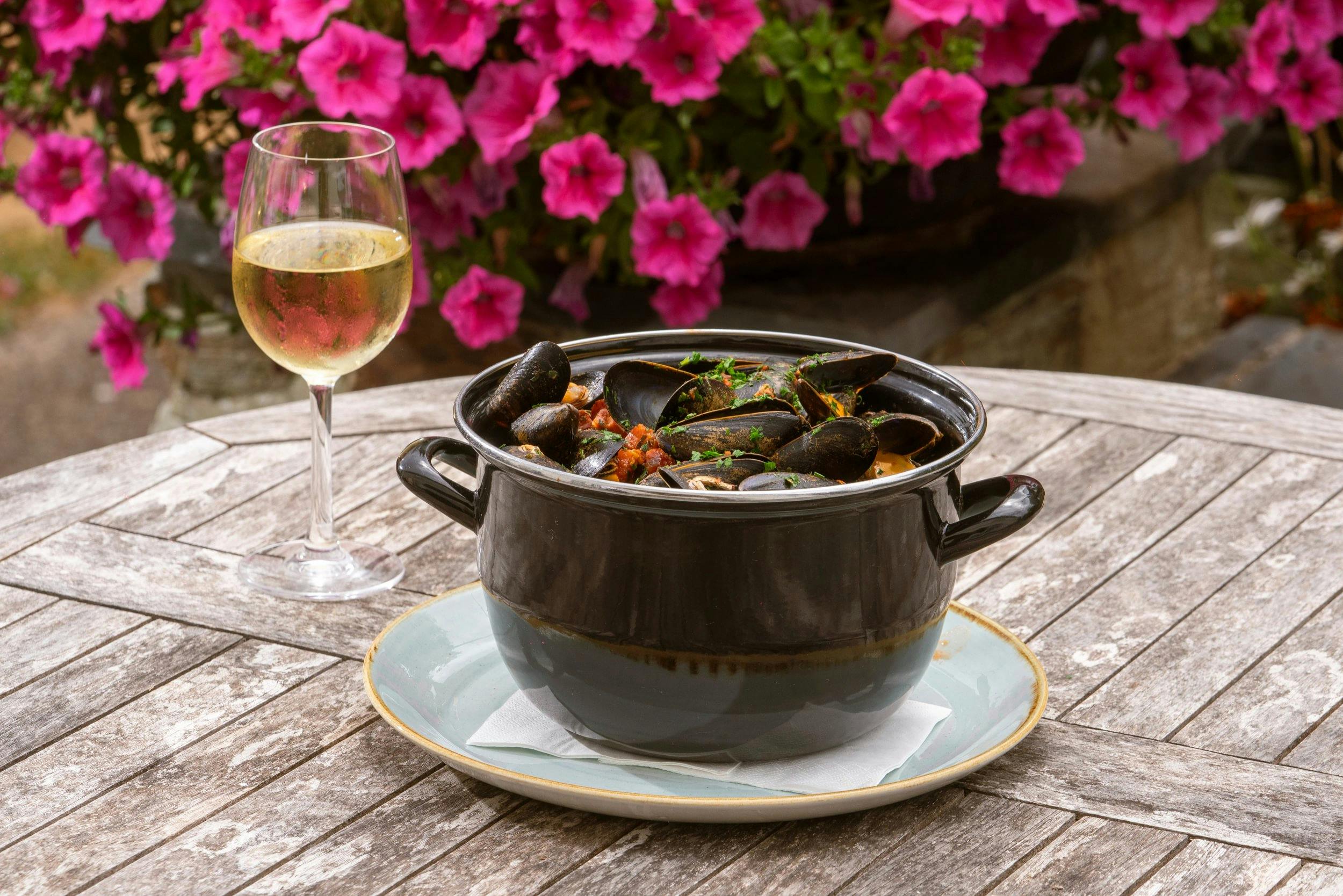 Black pot of mussels in tomato broth on a weathered outdoor table beside a glass of white wine, with bright pink flowers behind.