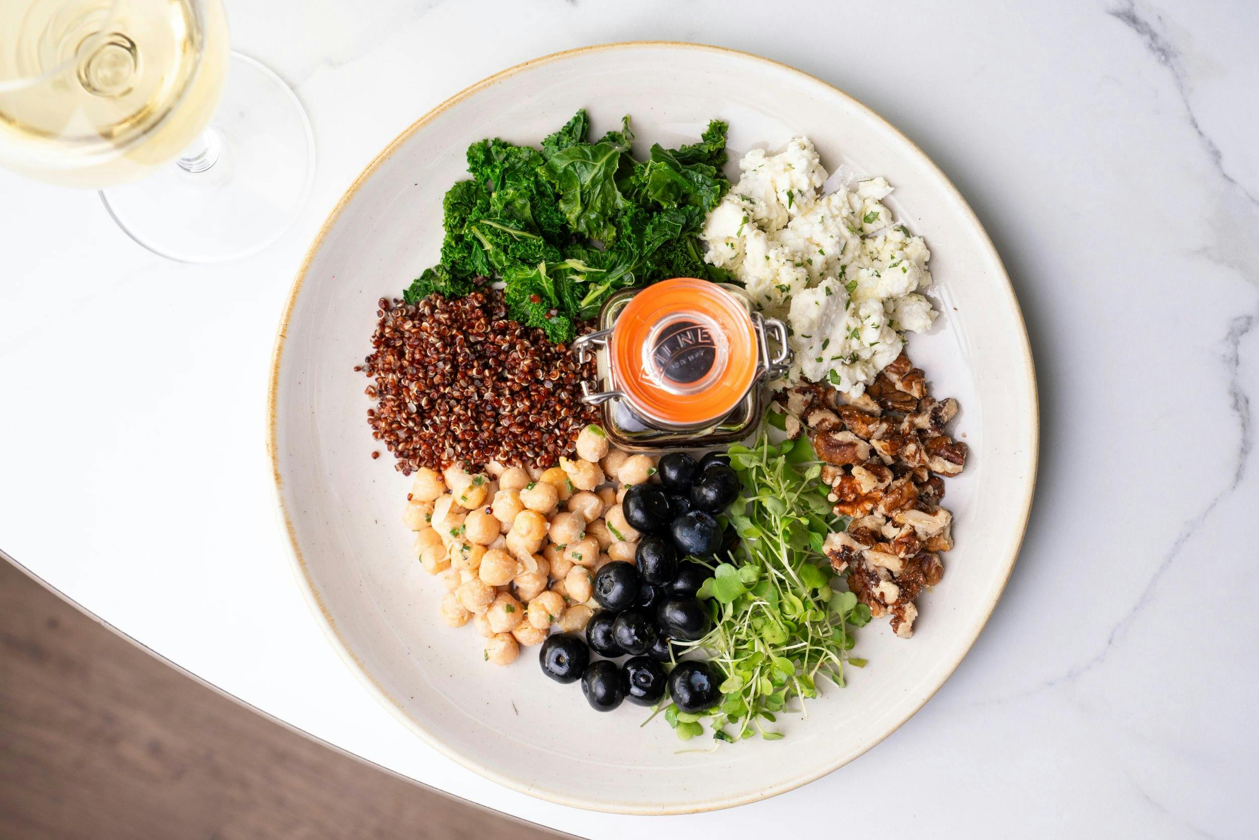 Overhead view of a grain bowl with kale, quinoa, chickpeas, olives, walnuts, microgreens, and cheese, plus a glass of white wine.