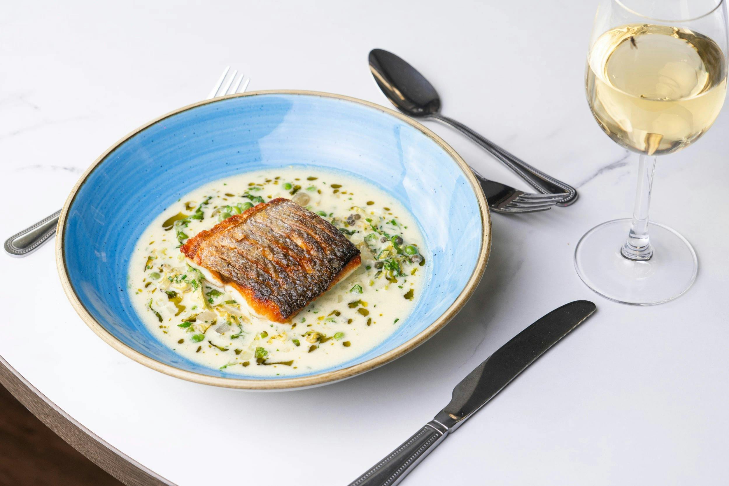 Crispy-skinned fish fillet in a creamy herb sauce in a blue bowl, set on a white table with cutlery and a glass of white wine.