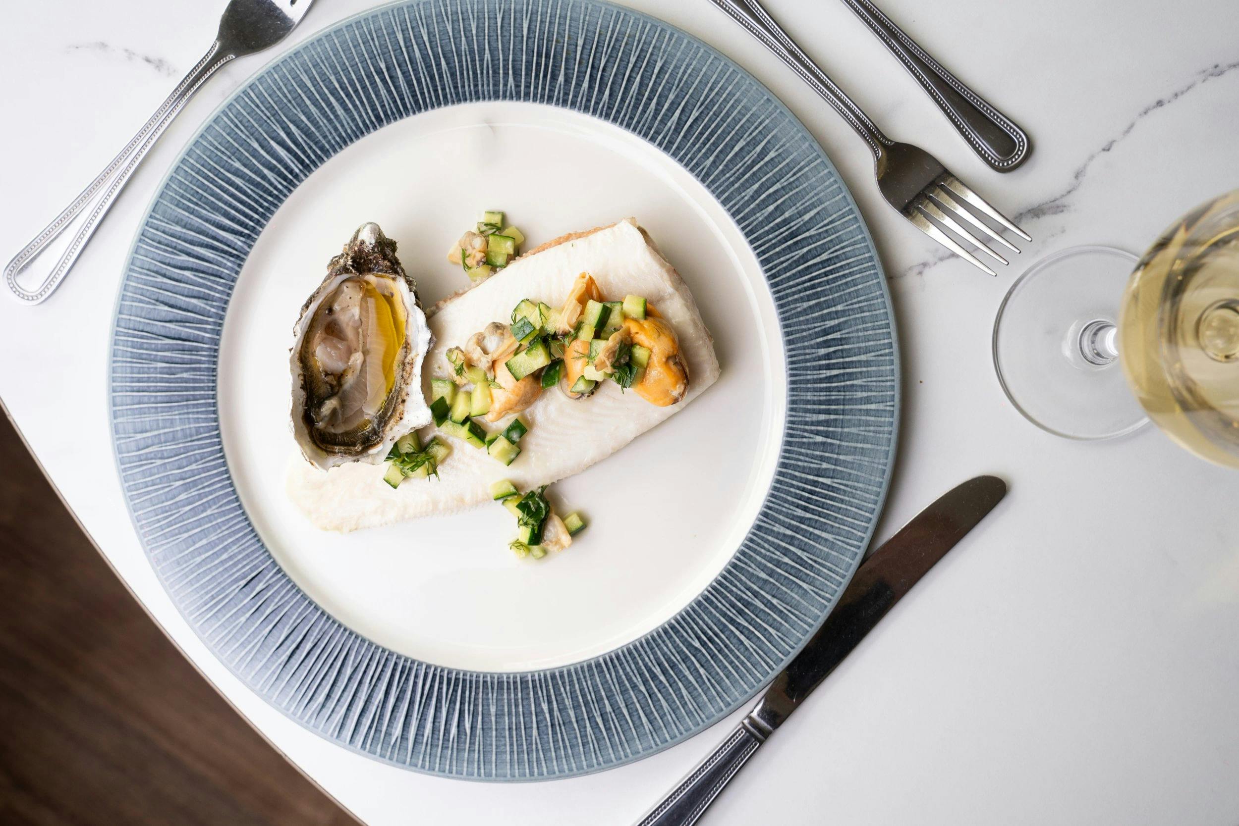 Overhead view of a plated white fish fillet topped with diced cucumber and shellfish, beside an oyster, with wine and silverware.