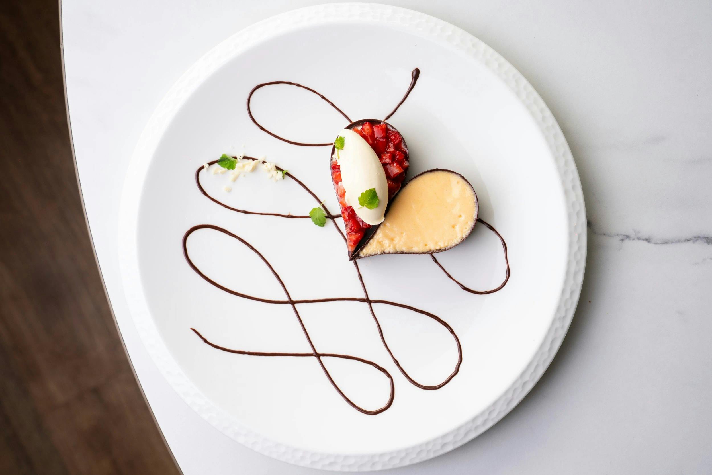 Plated dessert with a heart-shaped cheesecake, strawberry tart, vanilla quenelle, and looping chocolate sauce on a white plate.