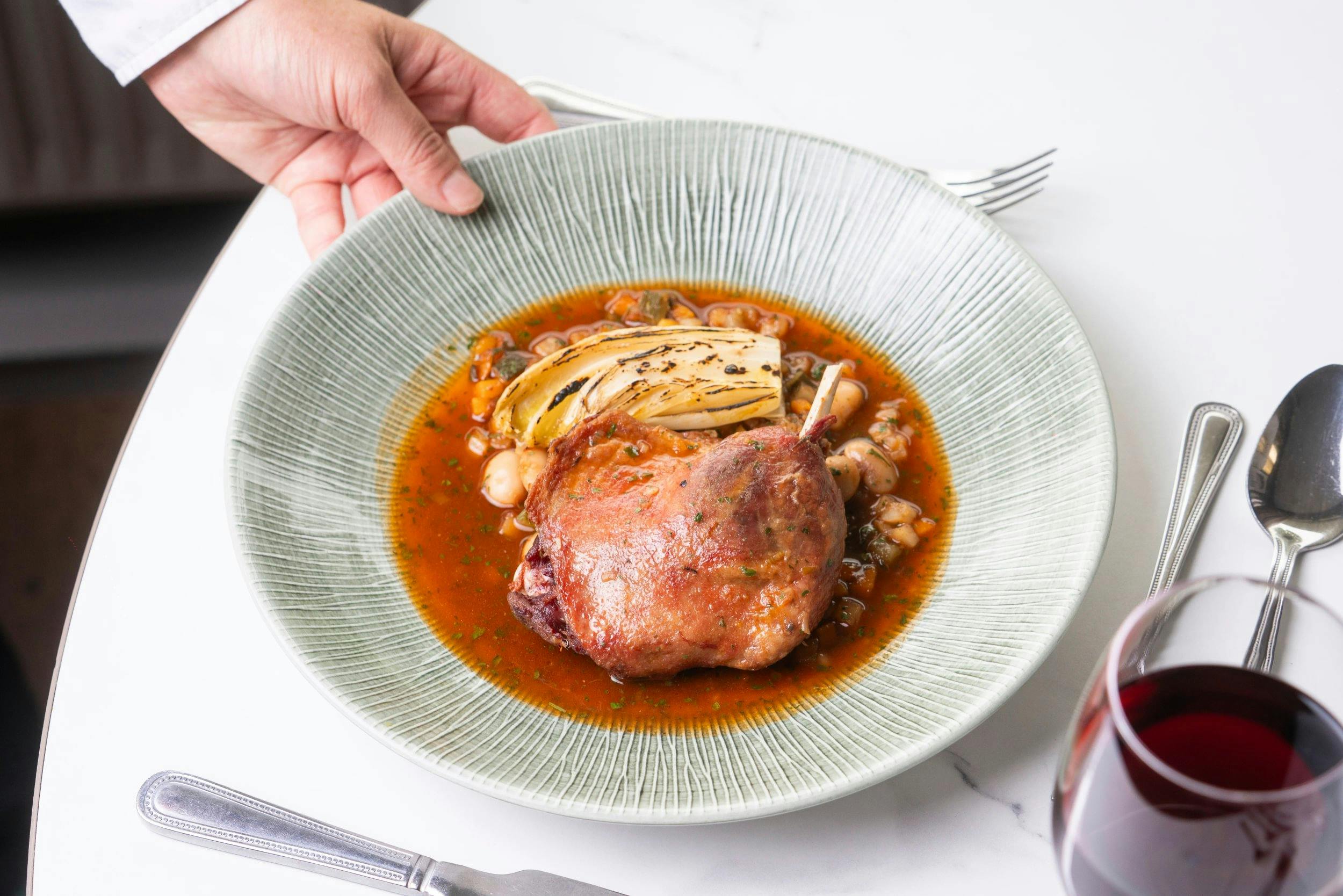 Roasted chicken leg in red broth with white beans and charred endive on a large grey plate, beside a glass of red wine.