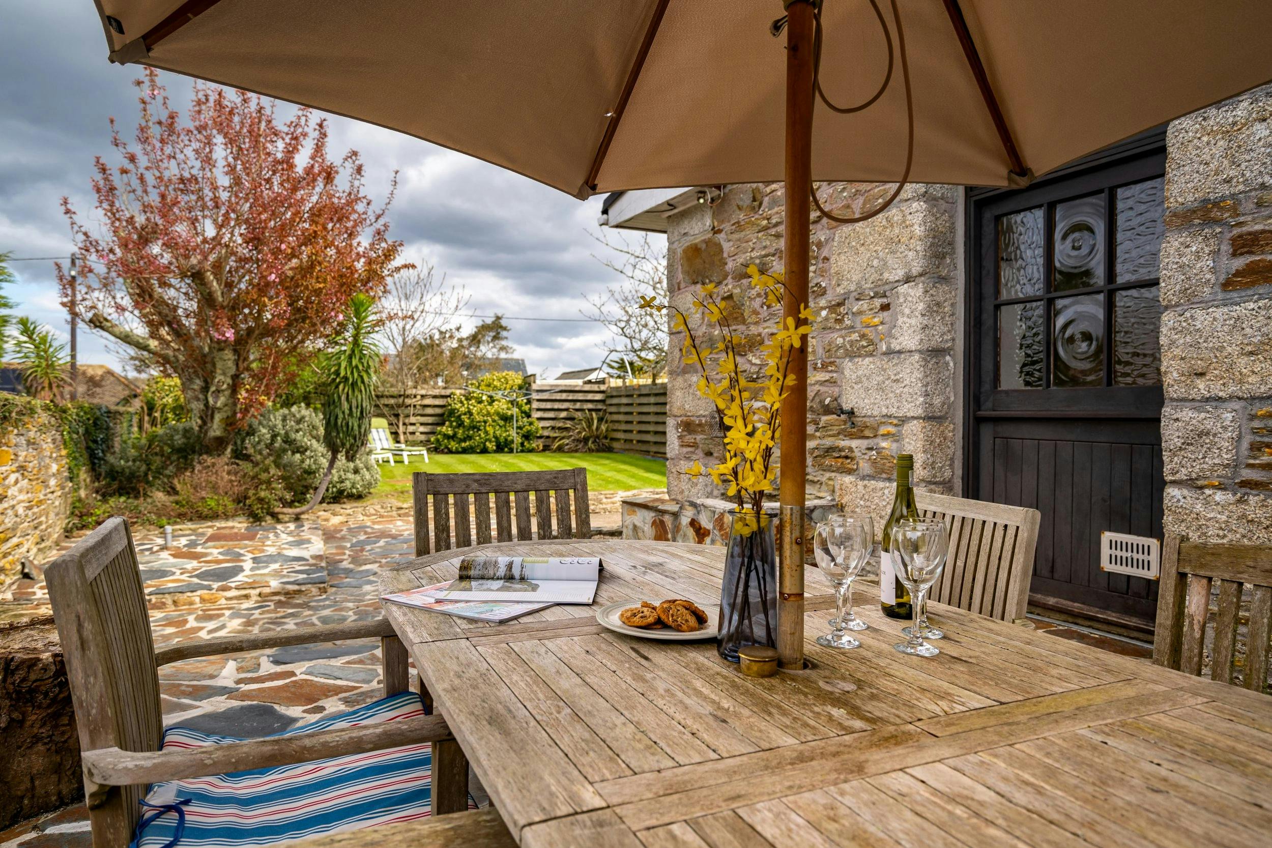 Patio table under an umbrella beside a stone cottage, set with wine, glasses, cookies, and flowers, overlooking a garden.
