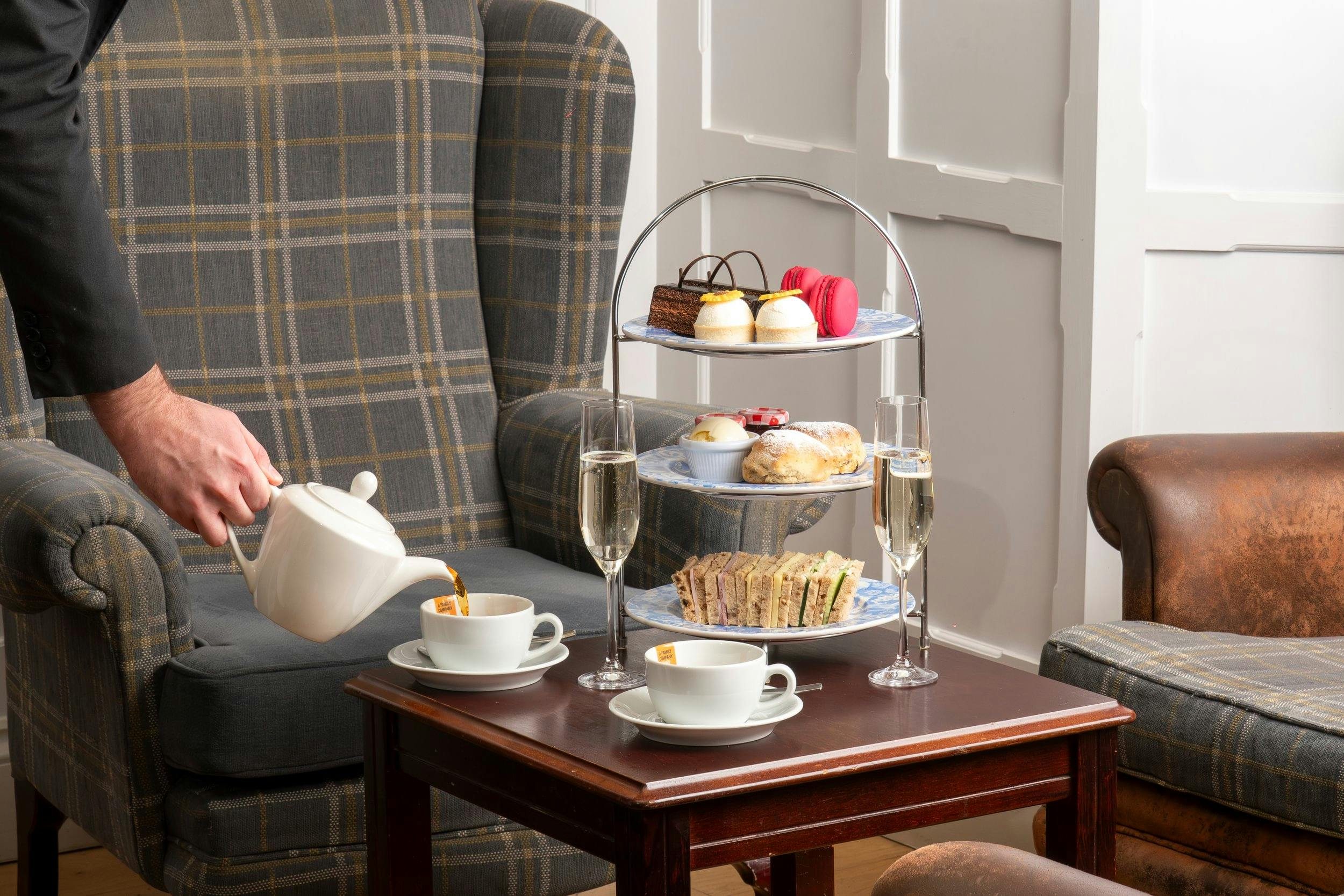 A person pours tea into a cup beside a tiered tray of pastries and sandwiches, with two glasses of champagne on a table.