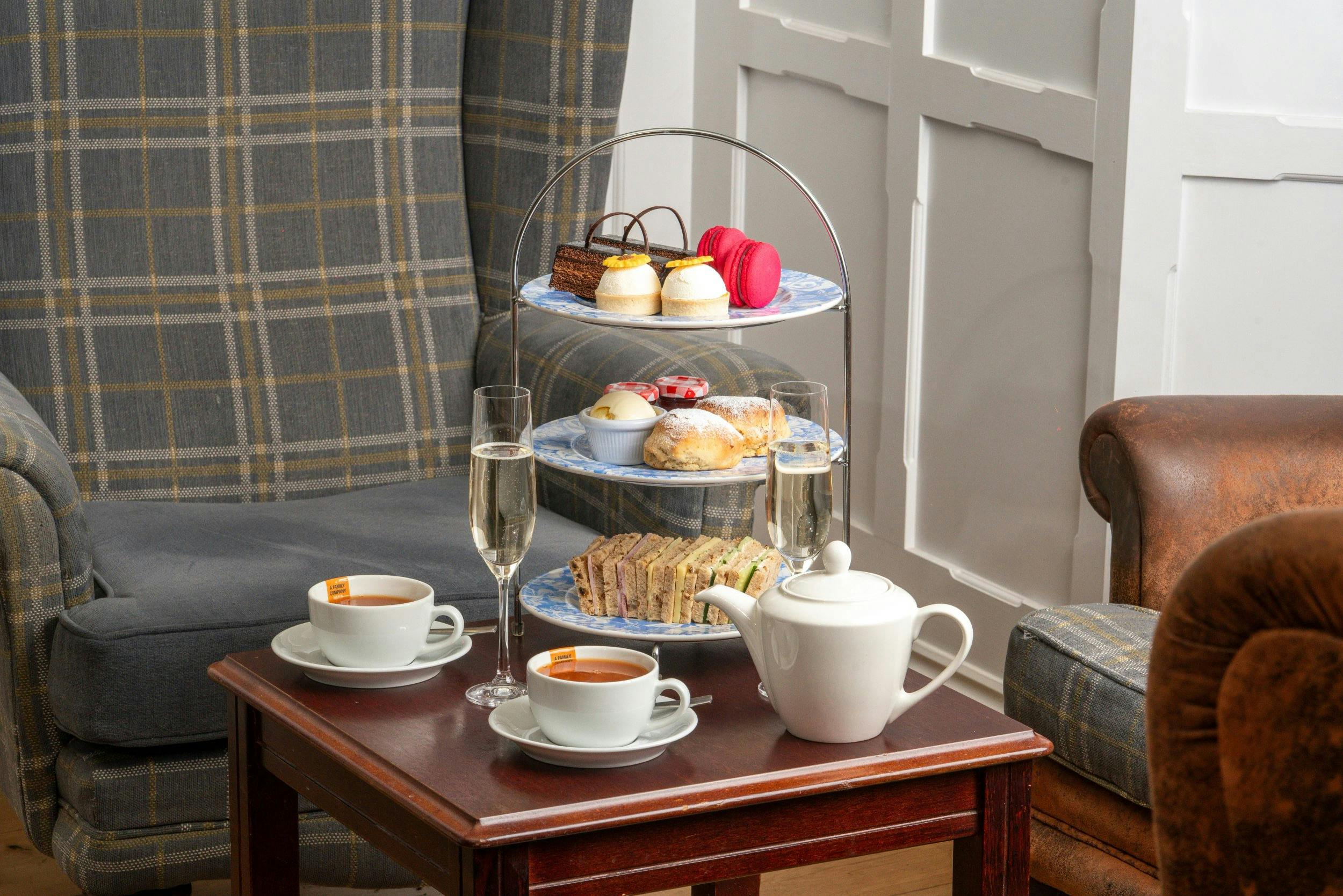 Afternoon tea on a wooden table with a tiered stand of sandwiches, scones, cakes, tea cups, teapot, and two flutes.