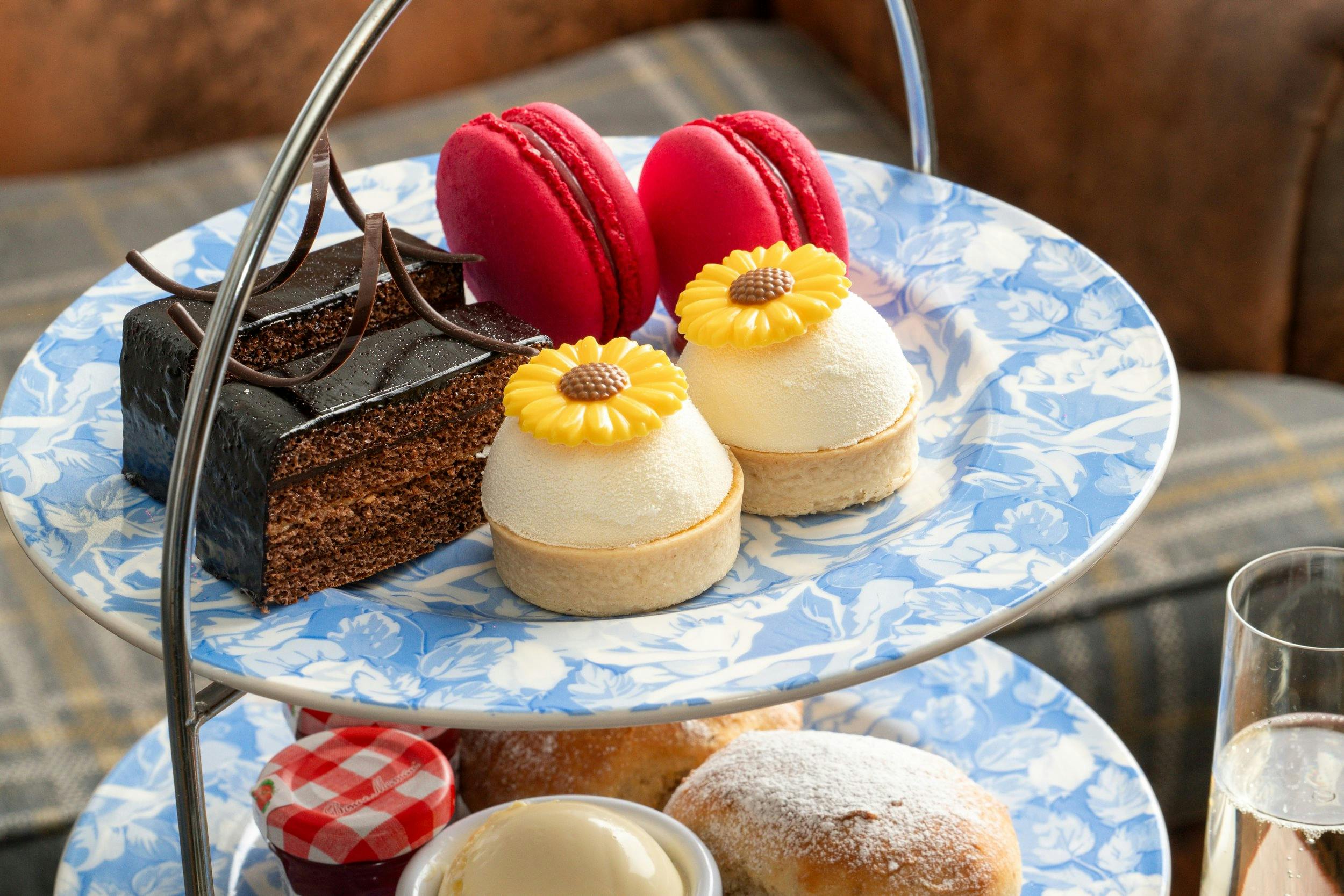 Tiered afternoon tea stand with chocolate cake slices, pink macarons, flower-topped mini tarts, scones, cream, and jam.