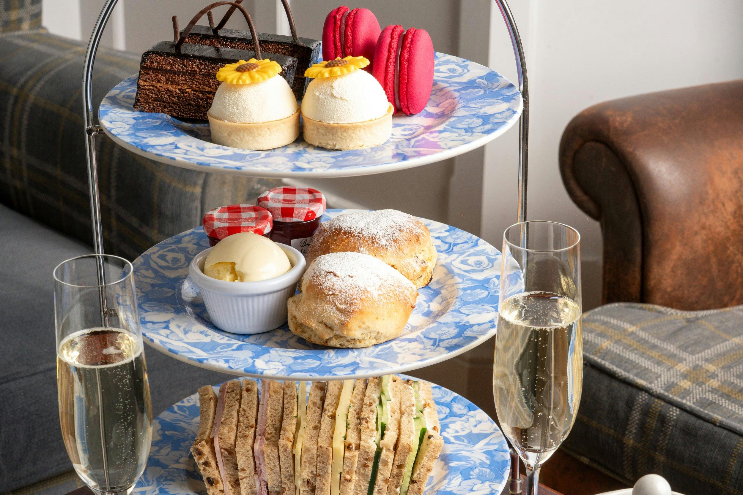 Three-tier afternoon tea stand with finger sandwiches, scones, cakes, pink macarons, and two glasses of sparkling wine.