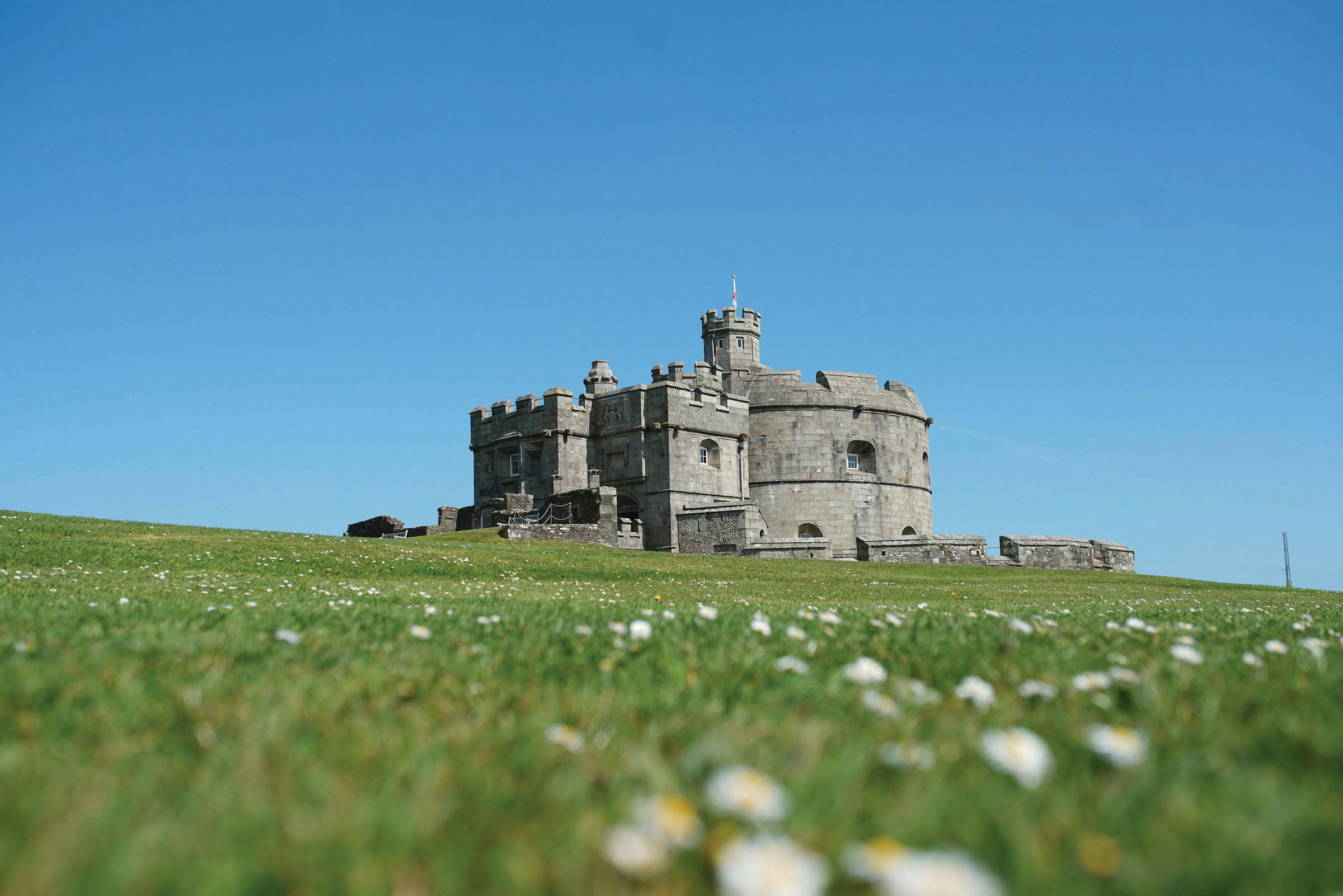 Stone castle-like building on a grassy hill dotted with white flowers beneath a wide, clear blue sky.