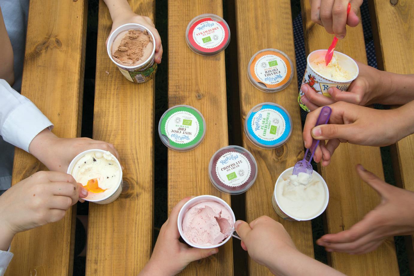 Several children sit around a wooden picnic table, eating cups of ice cream with colourful spoons; ice cream lids are scattered on the table.