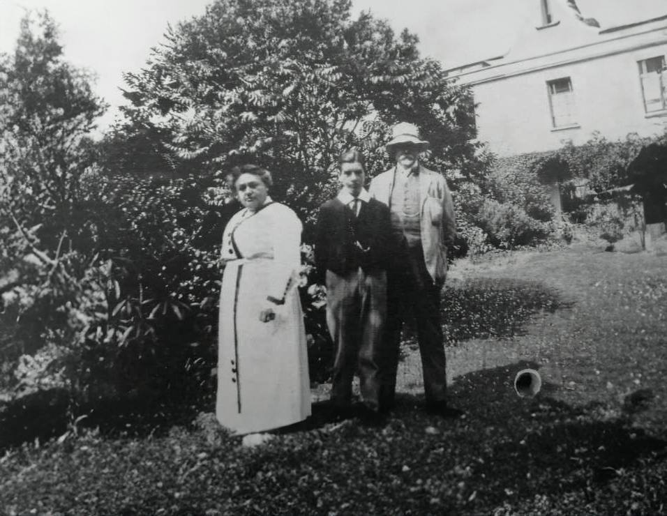 Black-and-white photo of a woman, a young man, and an older bearded man standing in a garden beside a house.