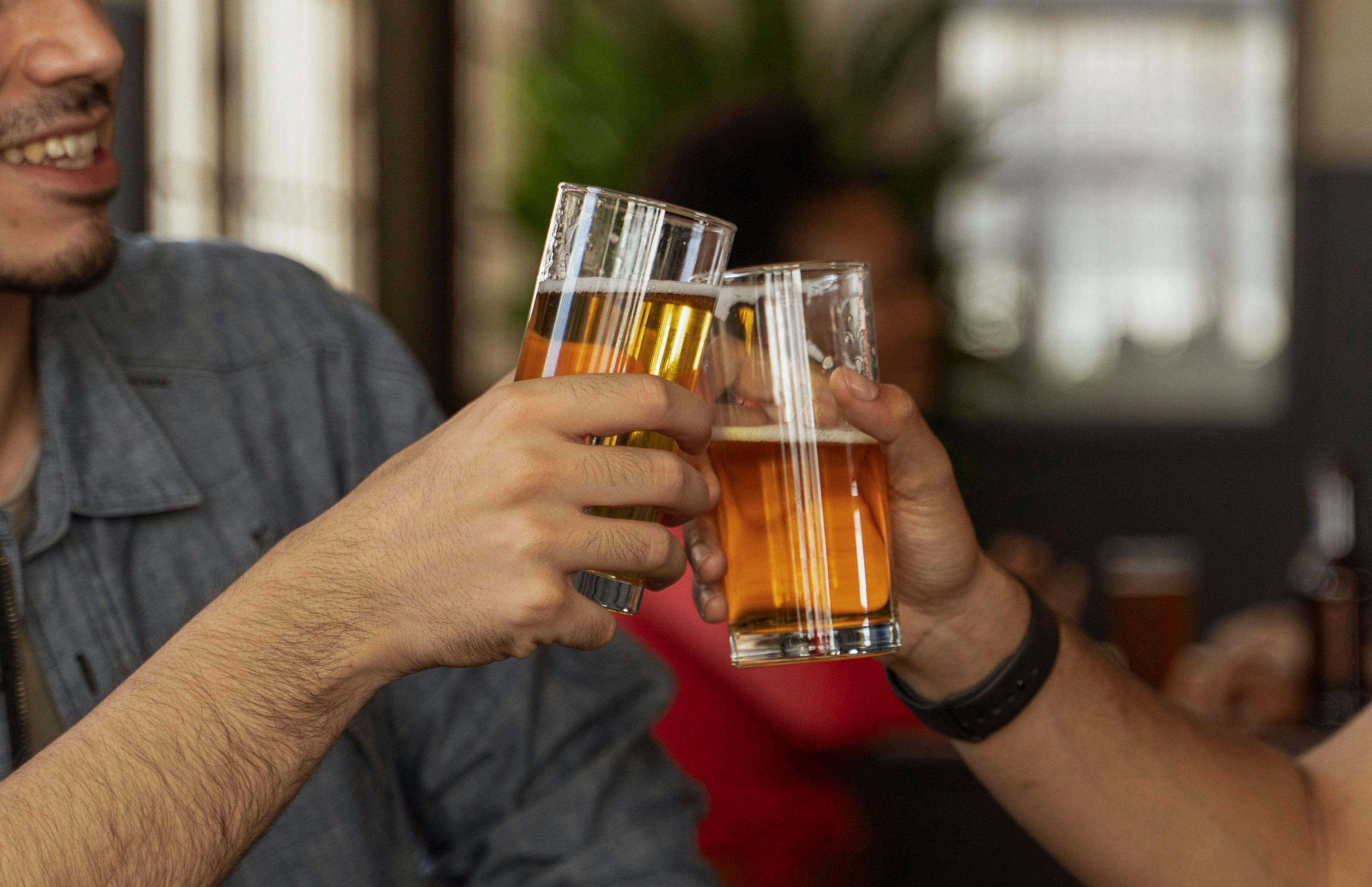 Two people clink tall glasses of amber beer in a bar, with one smiling face partially visible.
