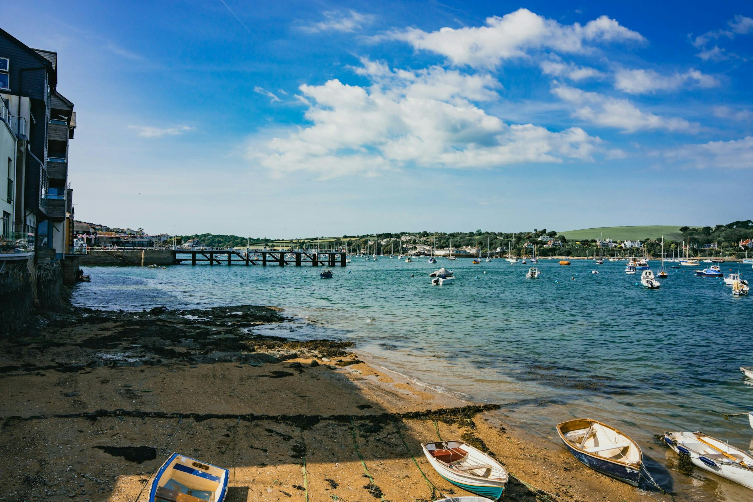 Sunny harbor scene with small boats on a sandy shore, a pier and waterfront buildings, and sailboats under a blue sky.