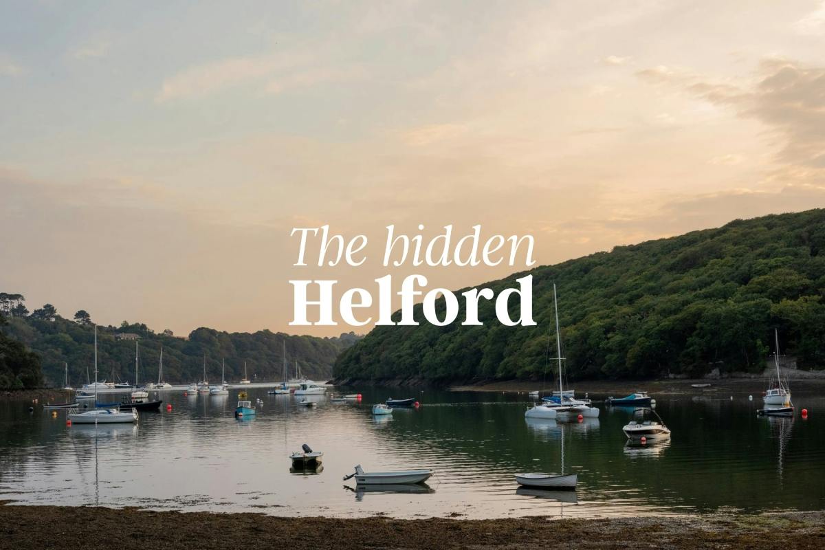 Calm river inlet at sunset with small boats moored on still water, wooded hills on both sides, and text reading “The hidden Helford.”