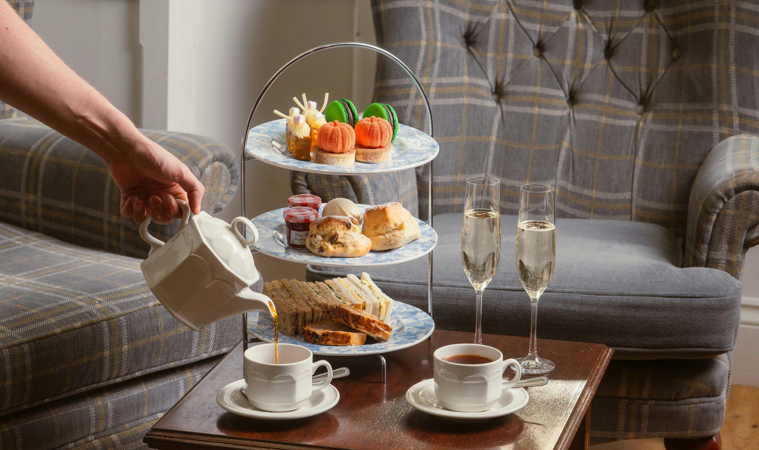 Hand pours tea beside a three-tier afternoon tea stand with sandwiches, scones, pastries, and two glasses of sparkling wine.
