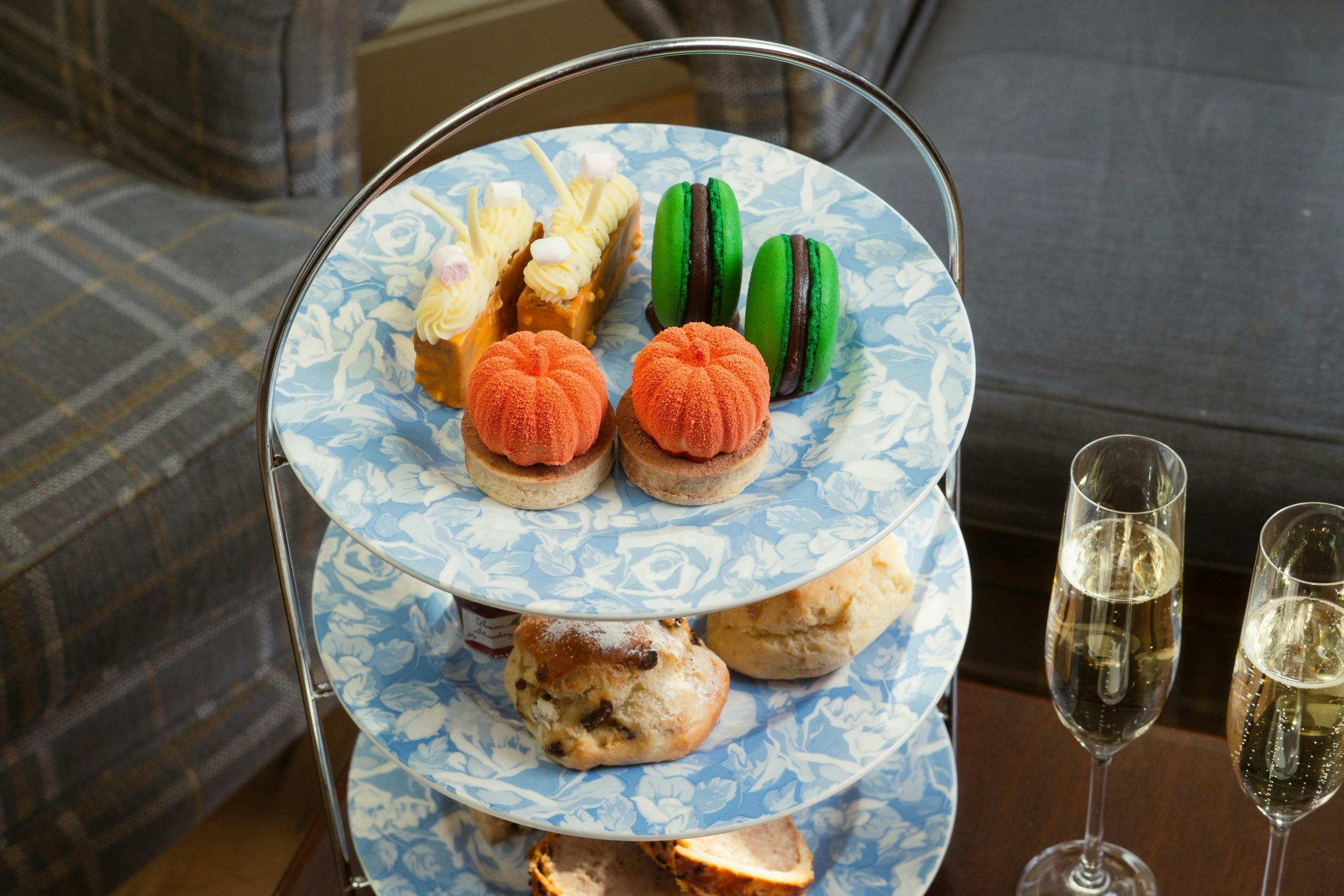 Three-tier afternoon tea stand with colourful pastries and scones beside two glasses of sparkling wine.