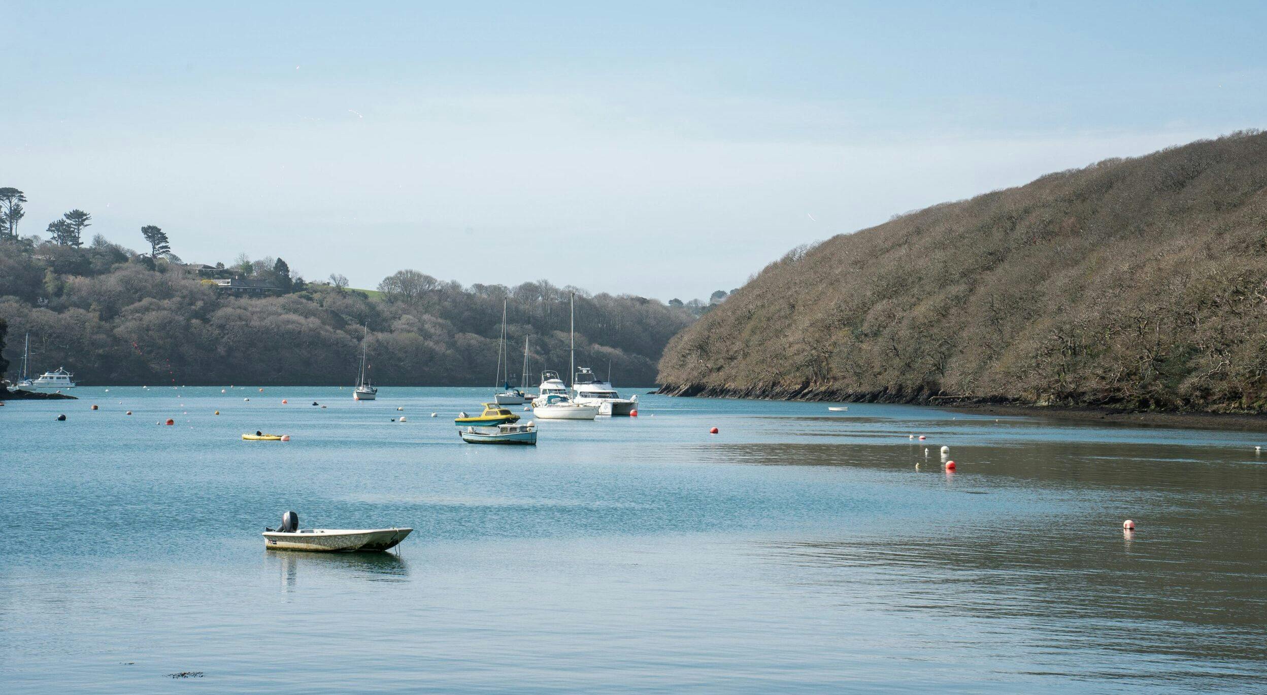 Calm estuary with small boats and buoys on blue water, bordered by wooded hills under a pale blue sky.
