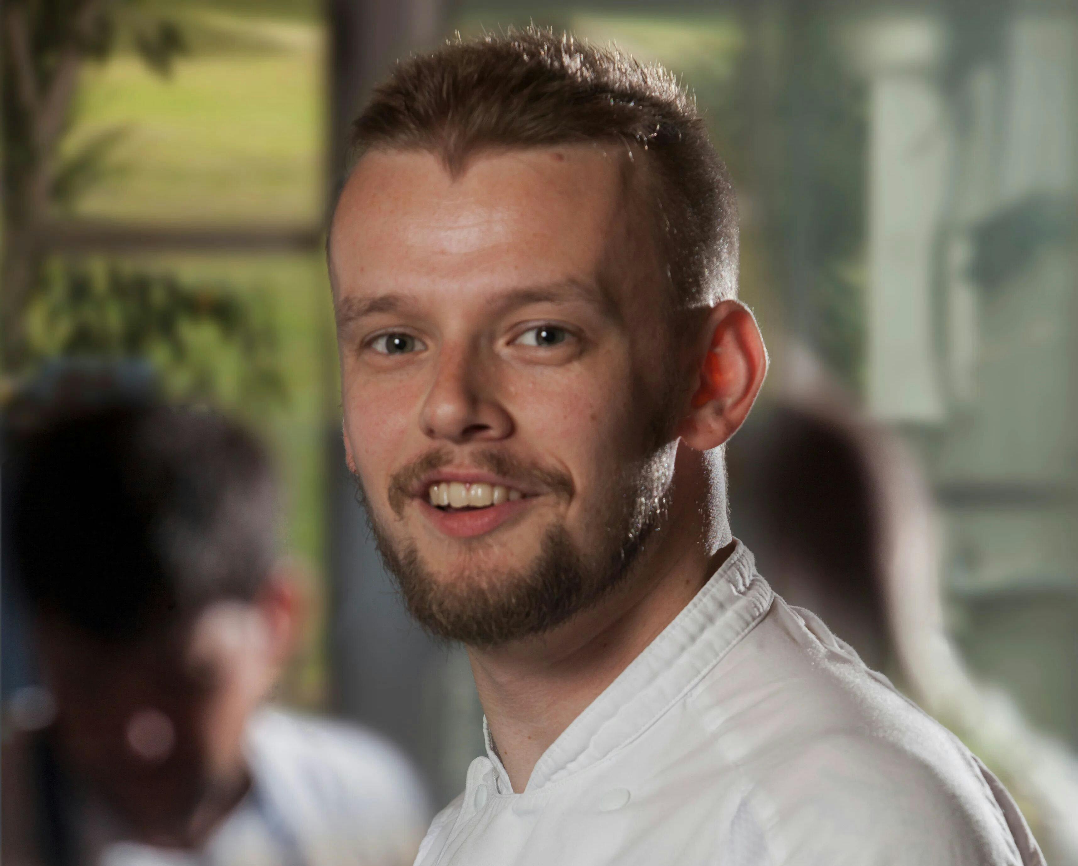 Smiling young man with short hair and a trimmed beard in a white chef’s jacket, indoors with blurred people in the background.