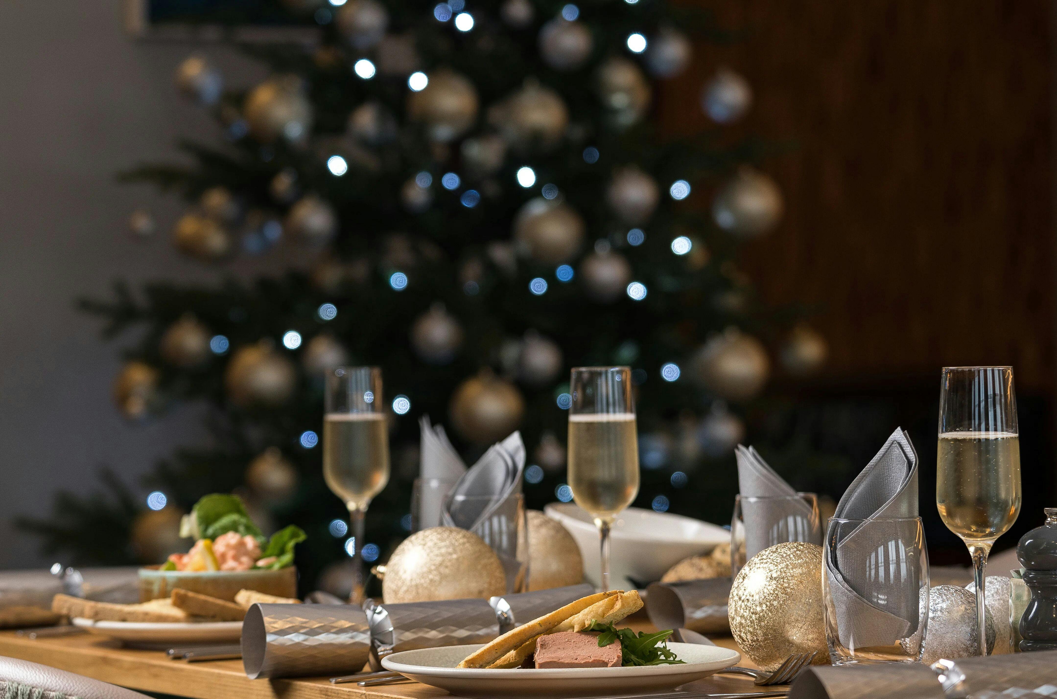 Festive holiday dinner table with champagne glasses and plates in front of a softly lit Christmas tree.