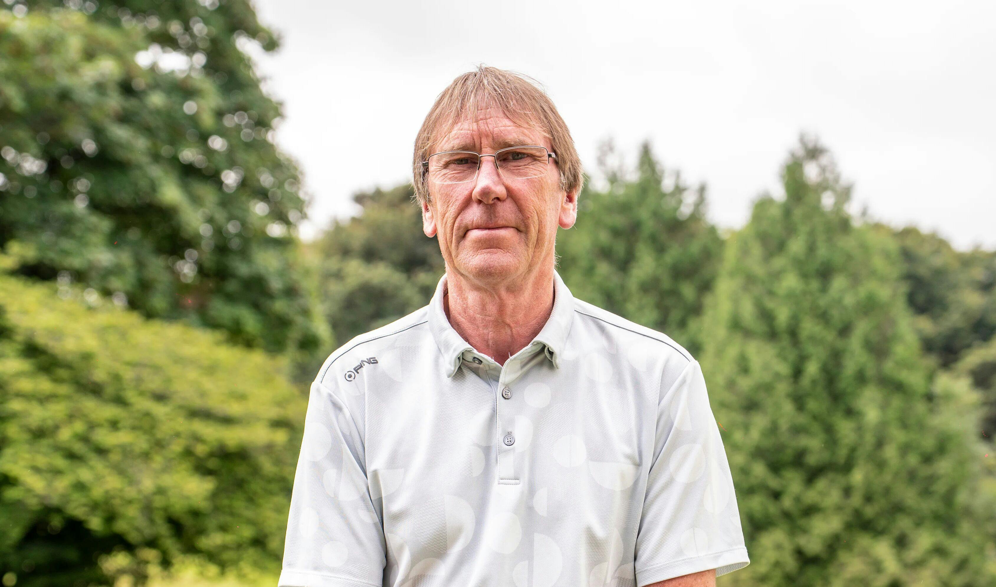Portrait of a man wearing glasses and a light polo shirt, standing outdoors with blurred green trees in the background.