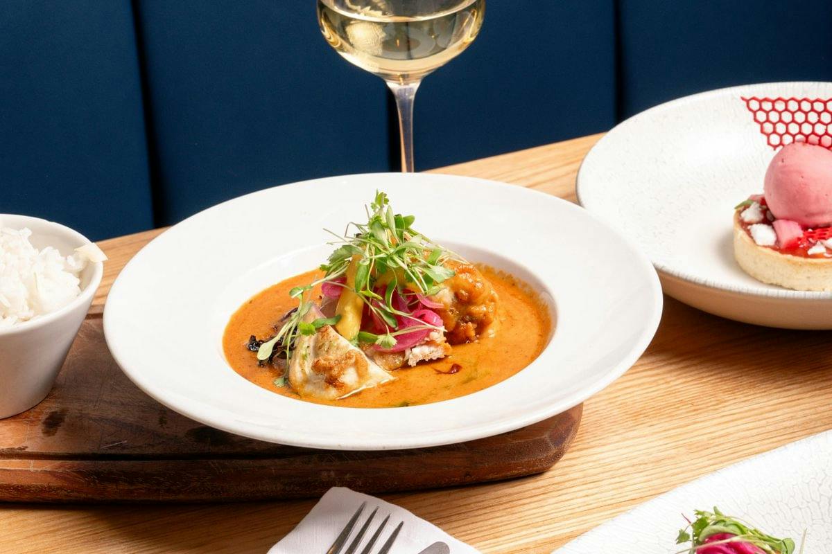 Plated curry with rice, a glass of white wine, and a pink dessert tart on a wooden table in a restaurant.