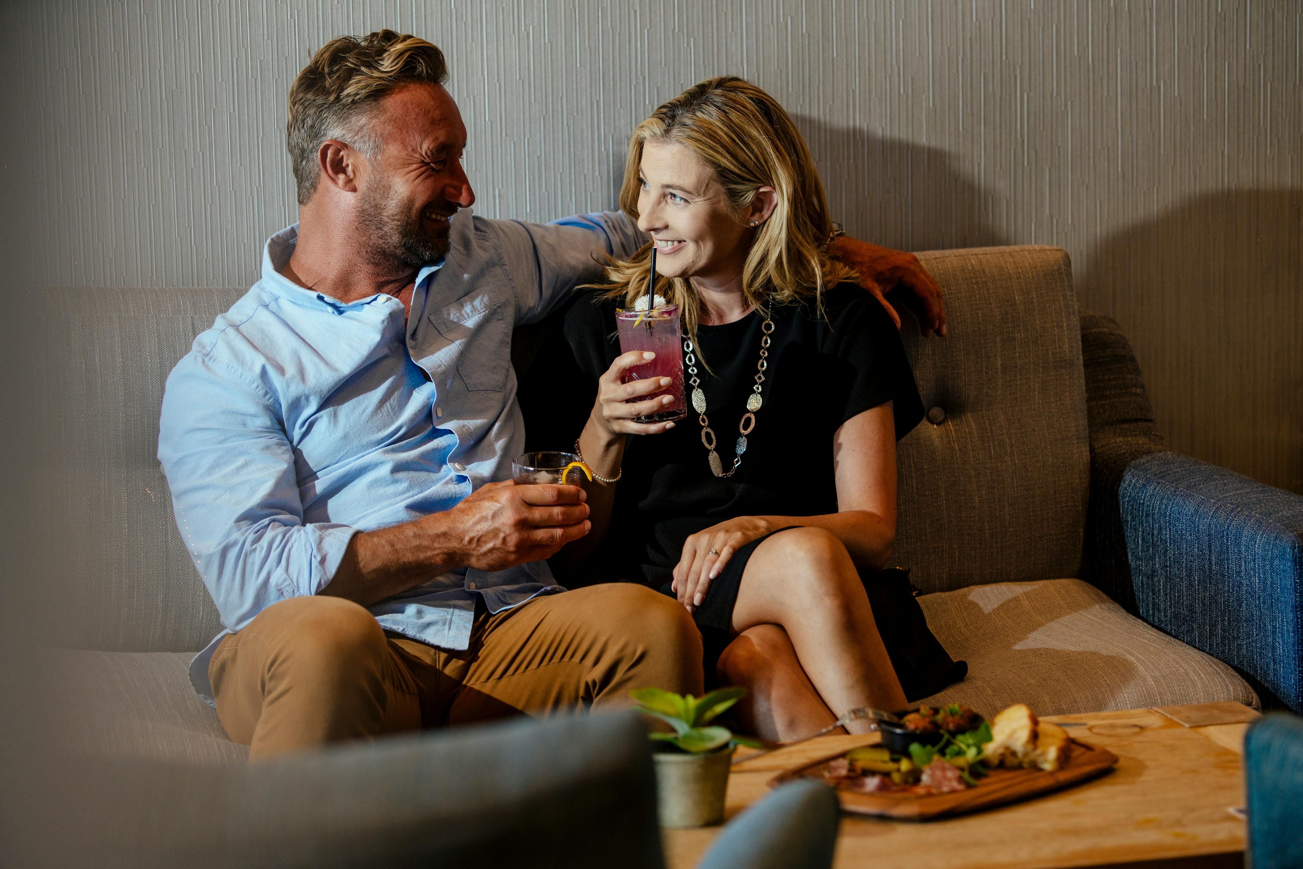 A couple sits on a sofa, smiling and holding drinks, with a small plant and food platter on the table in front of them.